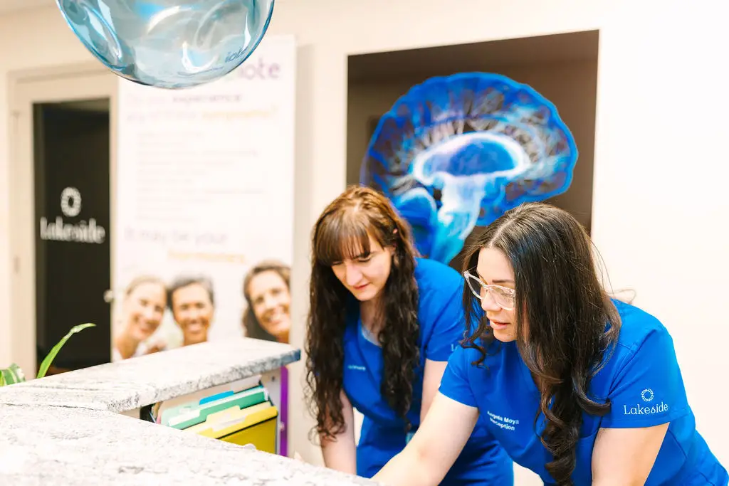 Two female staff in blue uniforms bend over a reception counter in a clinic, with a brain illustration on the wall behind them.
