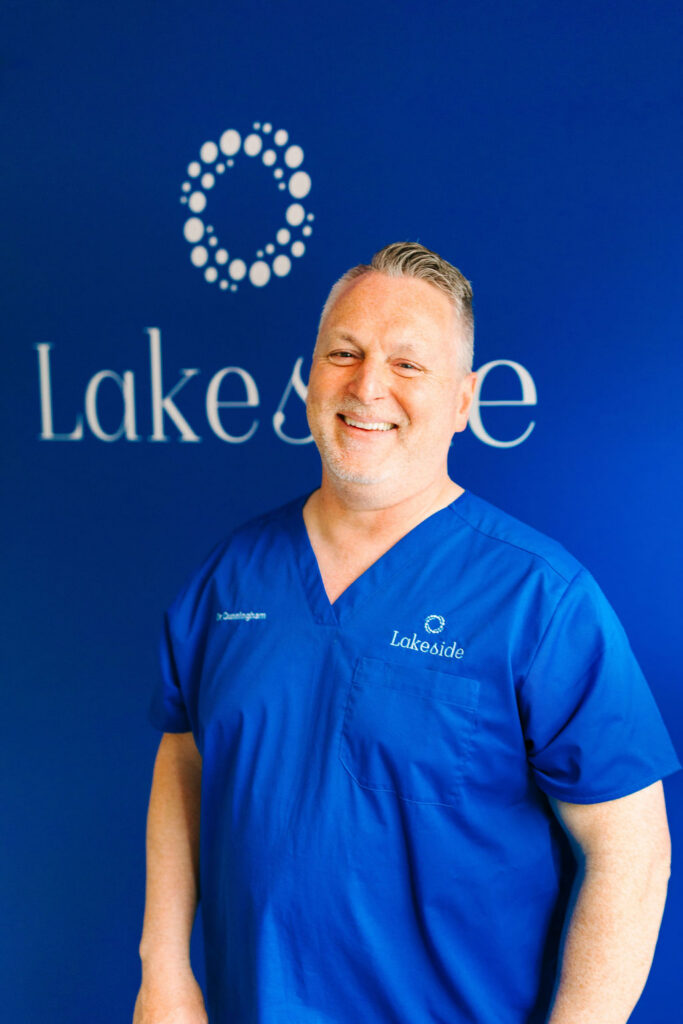 Smiling man in blue scrubs posing in front of a blue Lakeside backdrop with a circular logo pattern on the wall.