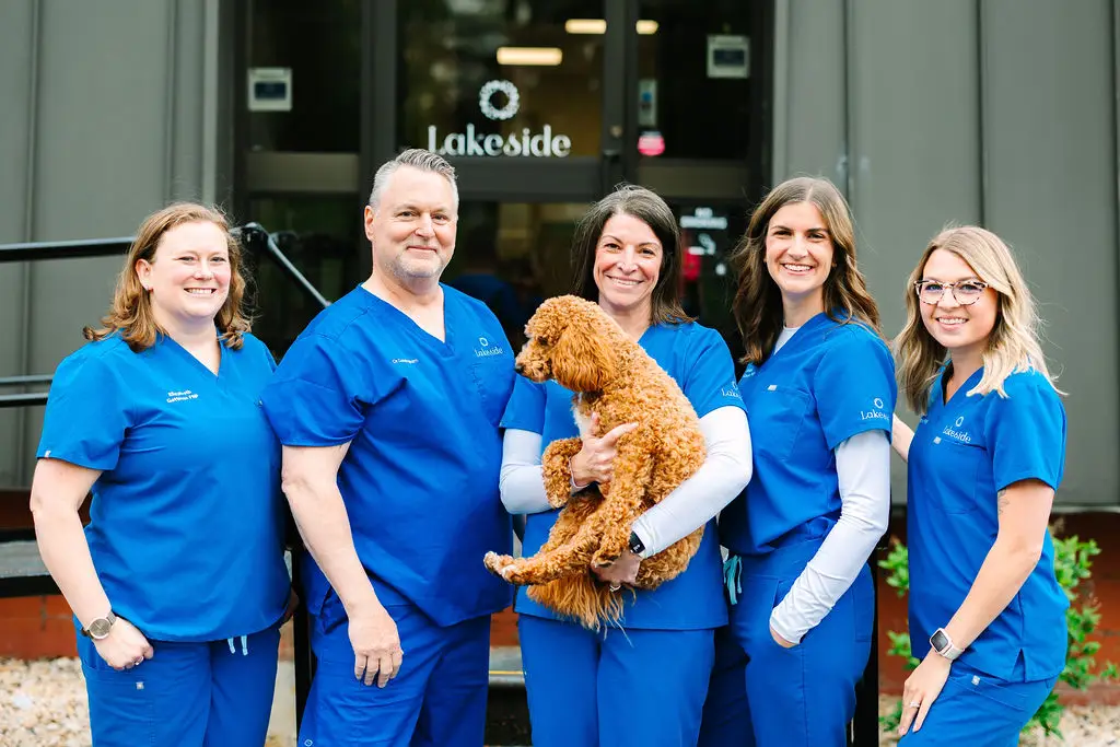 Five veterinary staff in blue scrubs stand outside a Lakeside building; one person holds a small brown poodle.